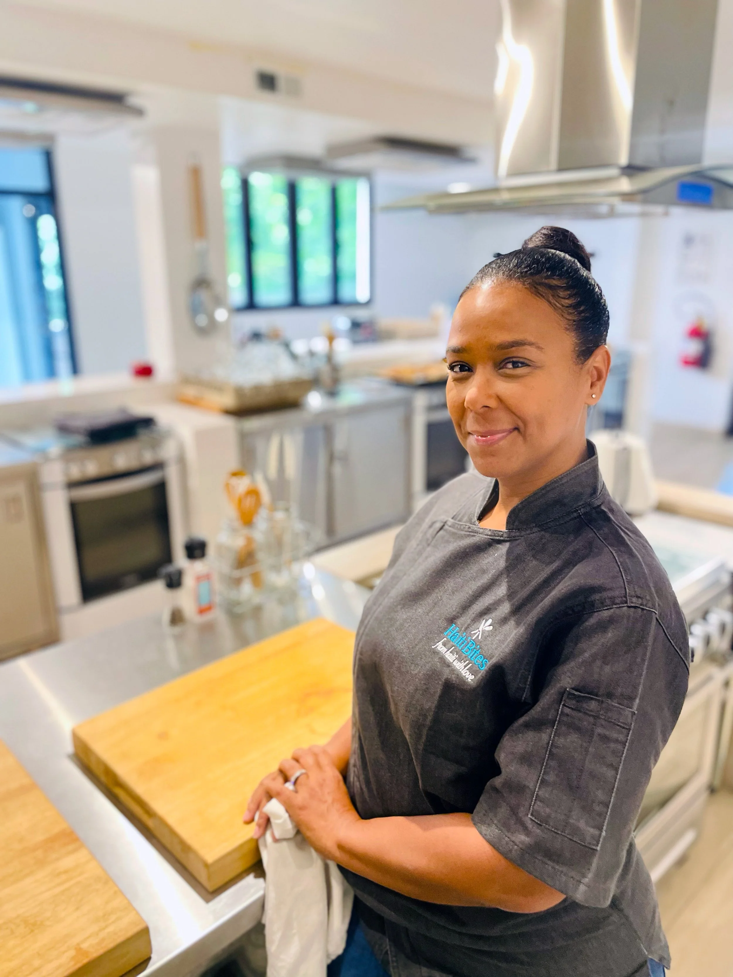 Chef standing in a kitchen with wooden cutting board, wearing a grey chef's jacket, smiling at the camera.