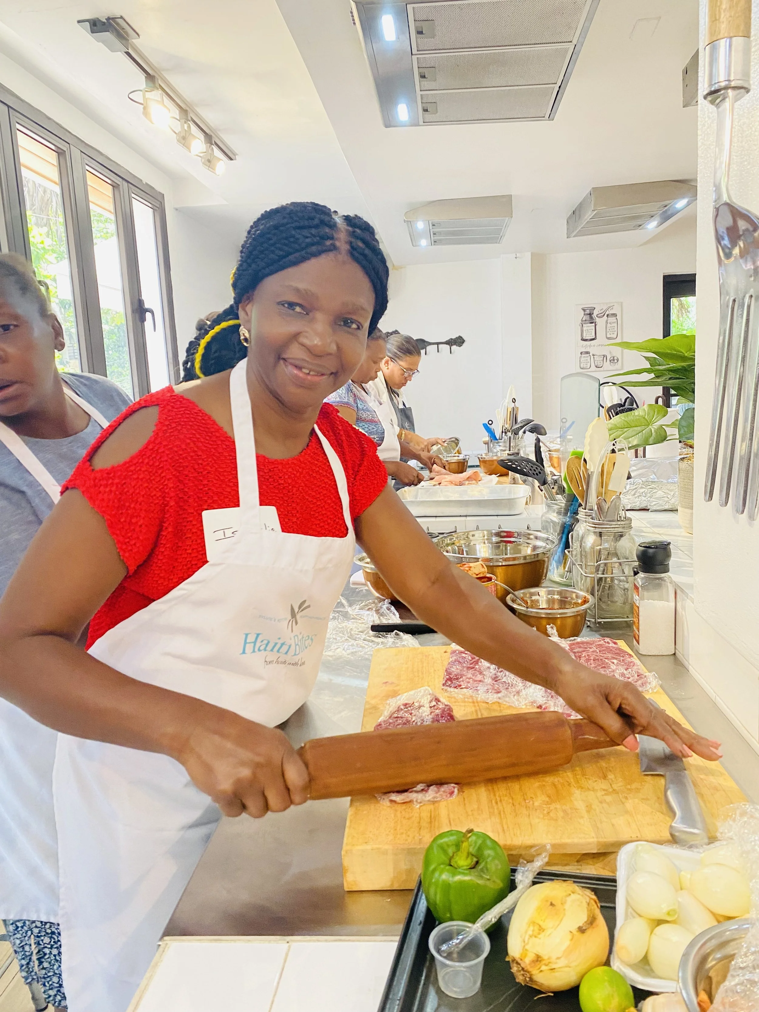 Woman in kitchen apron using rolling pin on meat, surrounded by vegetables like bell pepper and onions, in a cooking class setting.