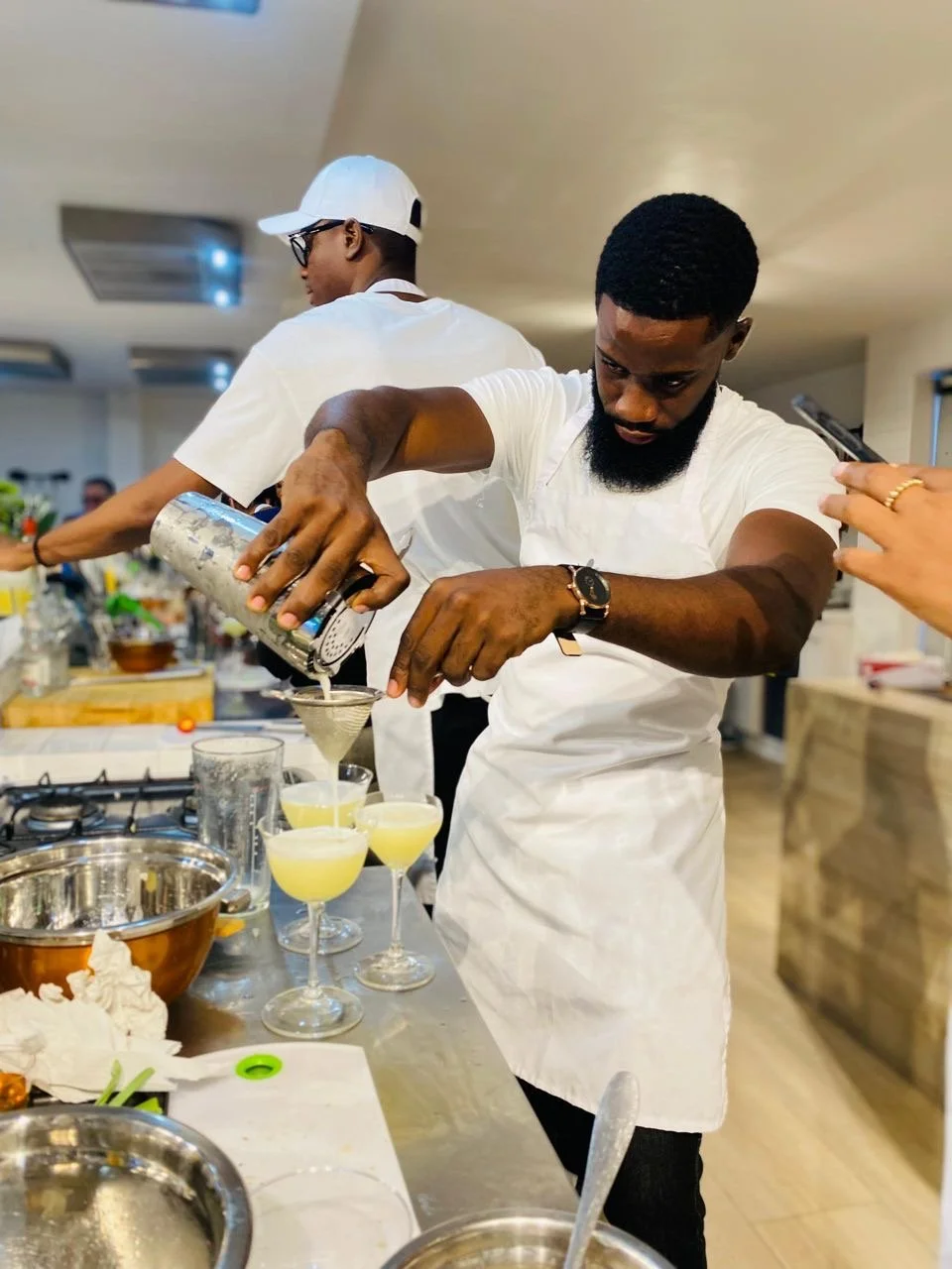 A bearded man in a white T-shirt and apron preparing cocktails in a kitchen. He is pouring a yellow drink through a strainer into a row of glasses. Another person wearing a white cap and glasses is seen in the background. The countertop has various kitchen utensils.