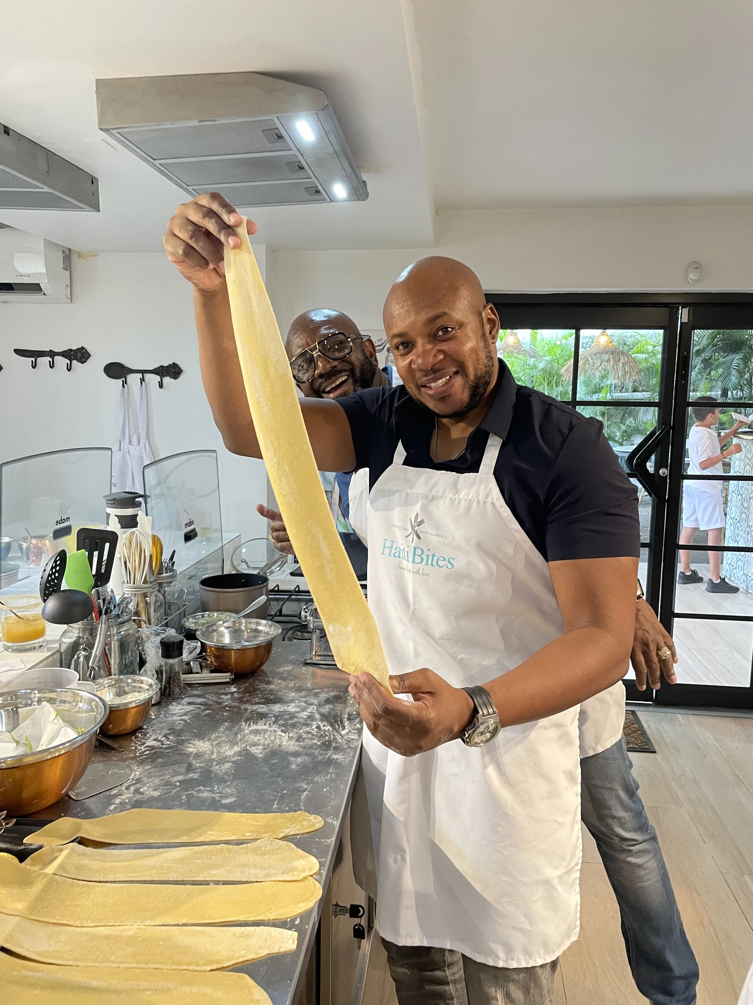 Two men wearing aprons making fresh pasta in a kitchen, one holding a long sheet of pasta dough.