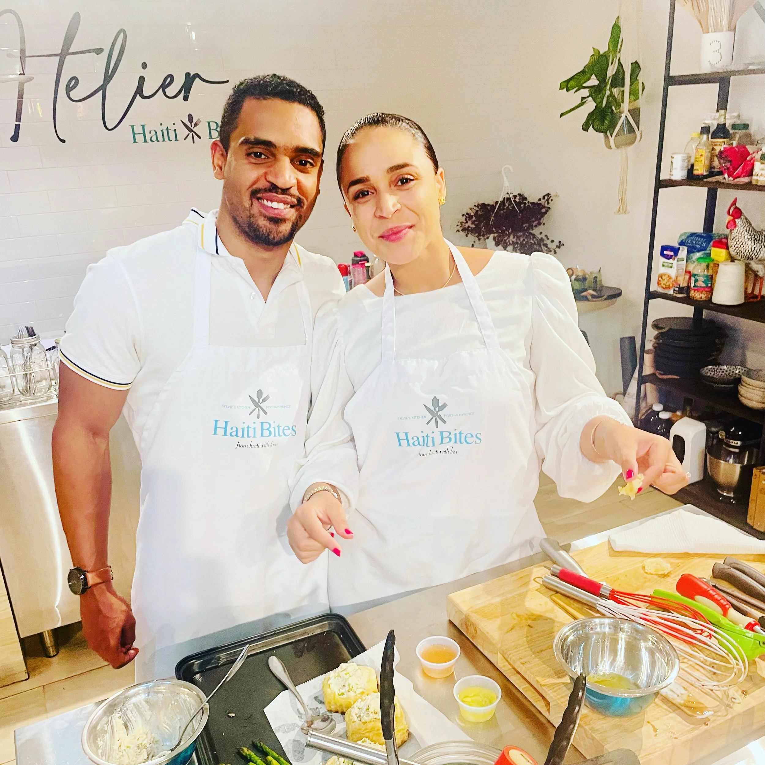 Two people wearing white aprons labeled "Haiti Bites" stand in a kitchen, smiling. They are beside cooking utensils, food preparations, and ingredients.