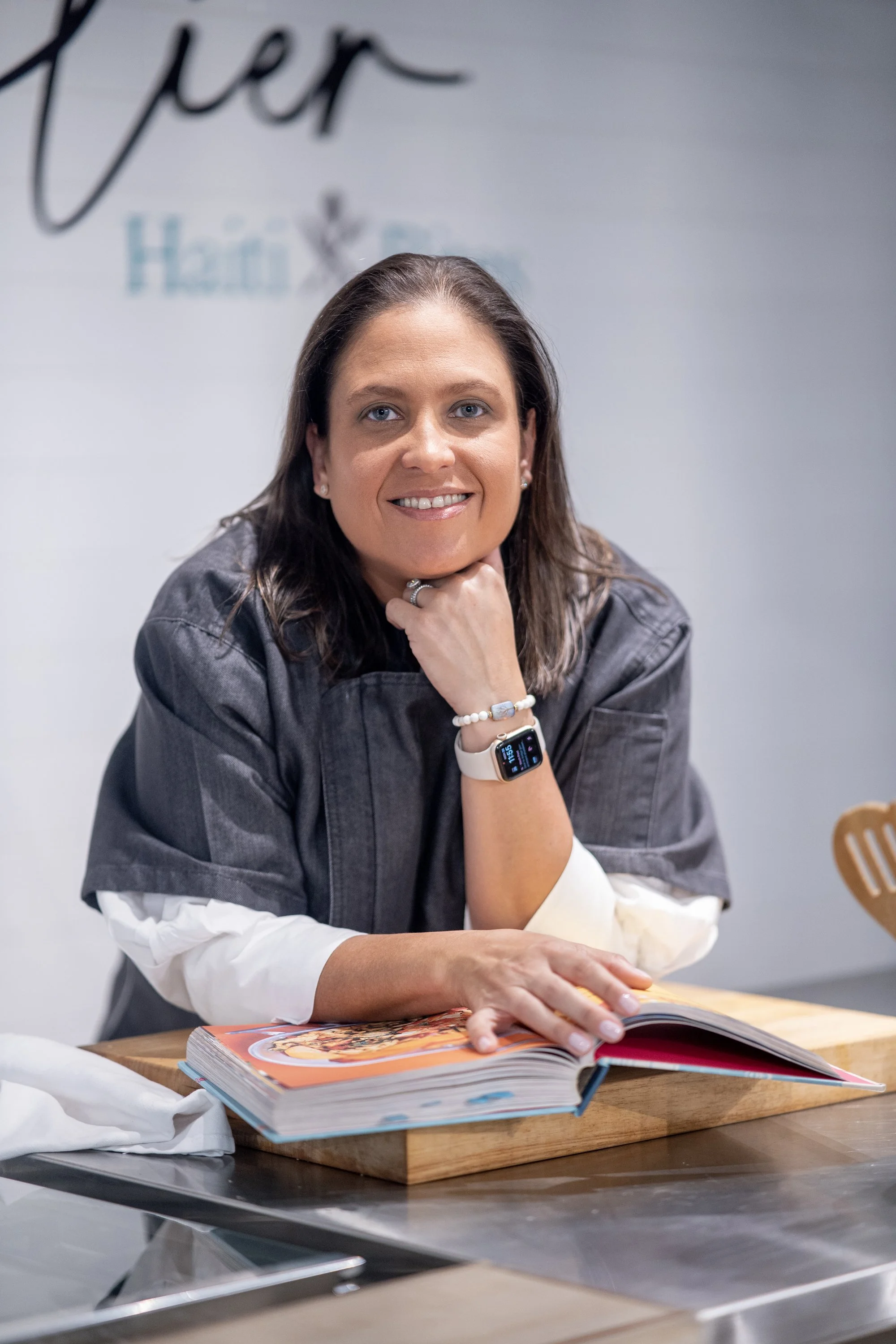 Chef Sylvie Castera Linge wearing a chef's jacket, smiling, with an open cookbook on the table.