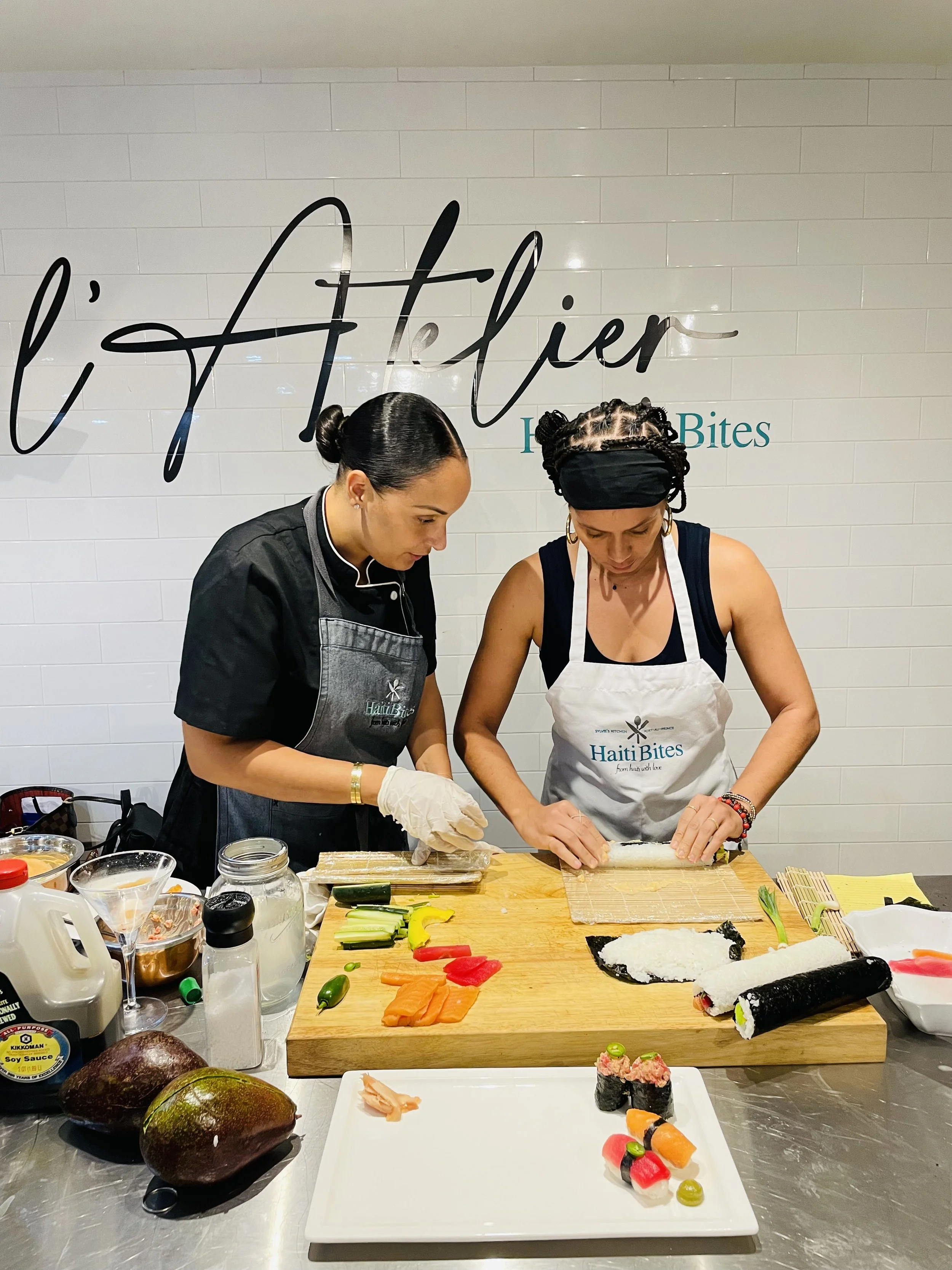 Two people making sushi rolls at a cooking class in L'Atelier with various ingredients like vegetables and sushi rice on a wooden cutting board.