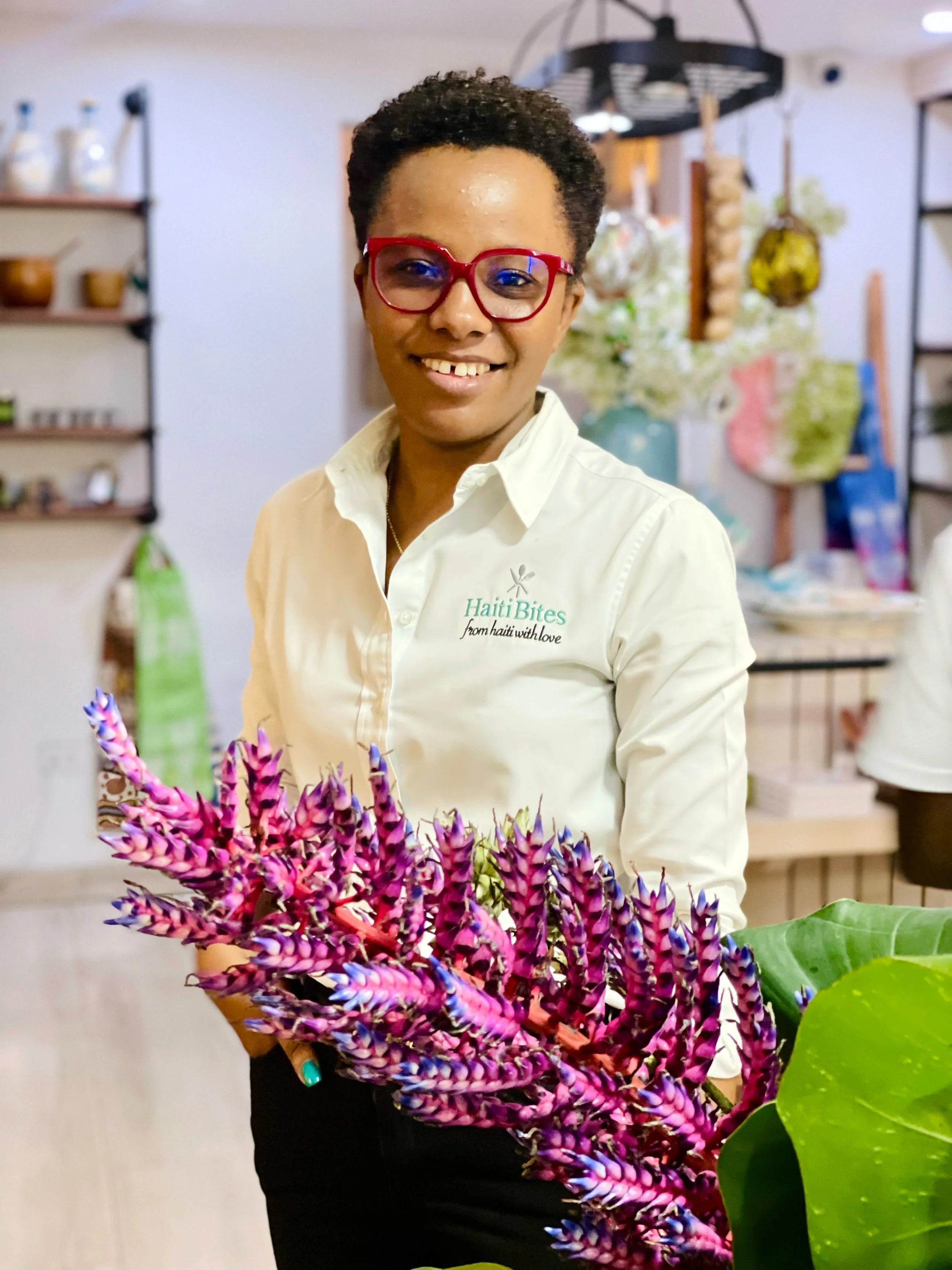 Person wearing glasses, in a white shirt with "HaitiBites" logo, holding a vibrant pink and purple tropical plant, standing indoors with shelves and hanging decor in the background.