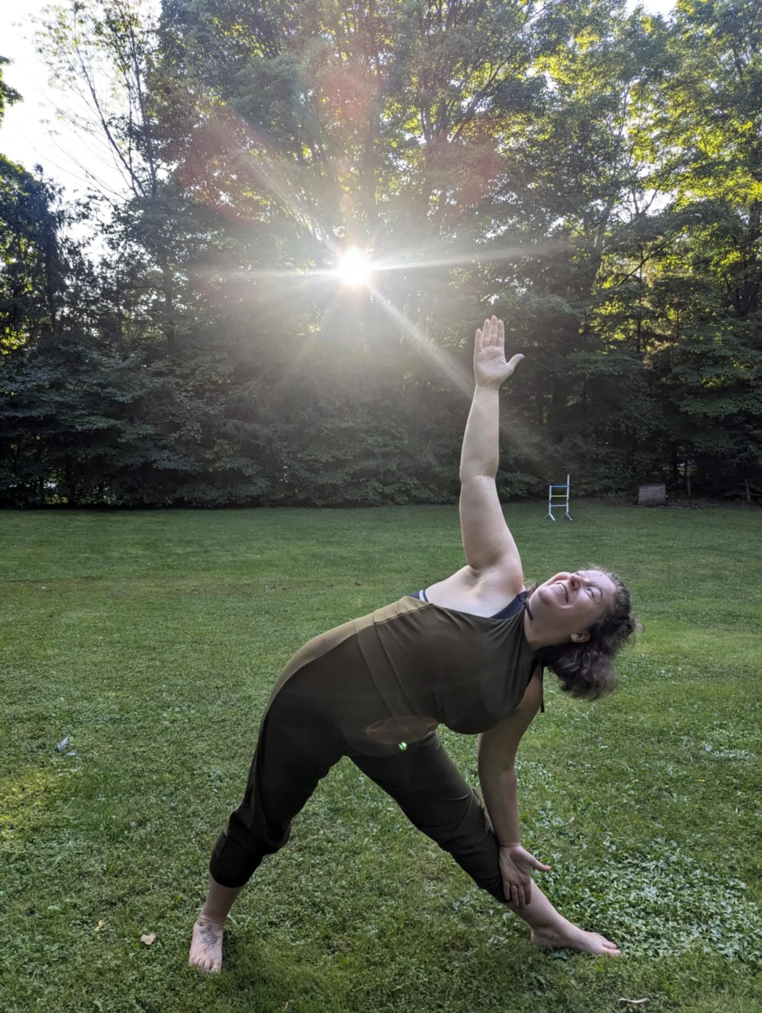 A person practicing yoga outdoors on grass, in a wide stance with one hand on the ground and the other stretched upward, smiling with a background of trees and sunlight.