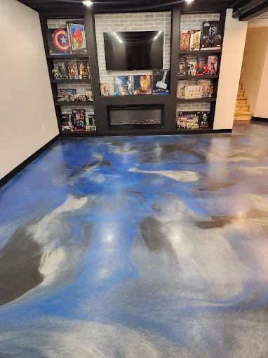 Living room with a black entertainment center and a mounted flat-screen TV, surrounded by shelves filled with collectibles and artwork, with a polished concrete floor featuring a swirling blue, gray, and white pattern.