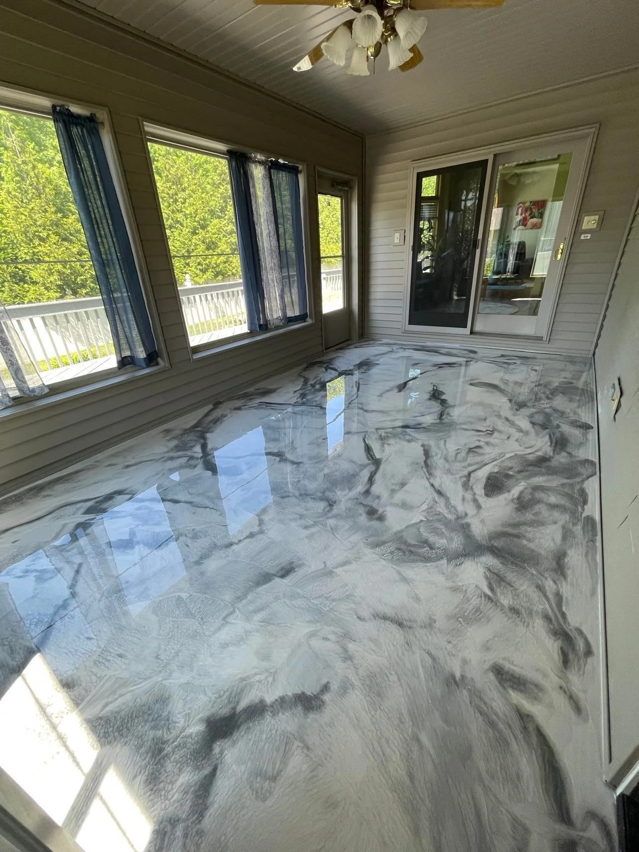 A room with large windows, a glass sliding door, and a ceiling fan, featuring a marble-patterned metallic floor with a glossy finish.