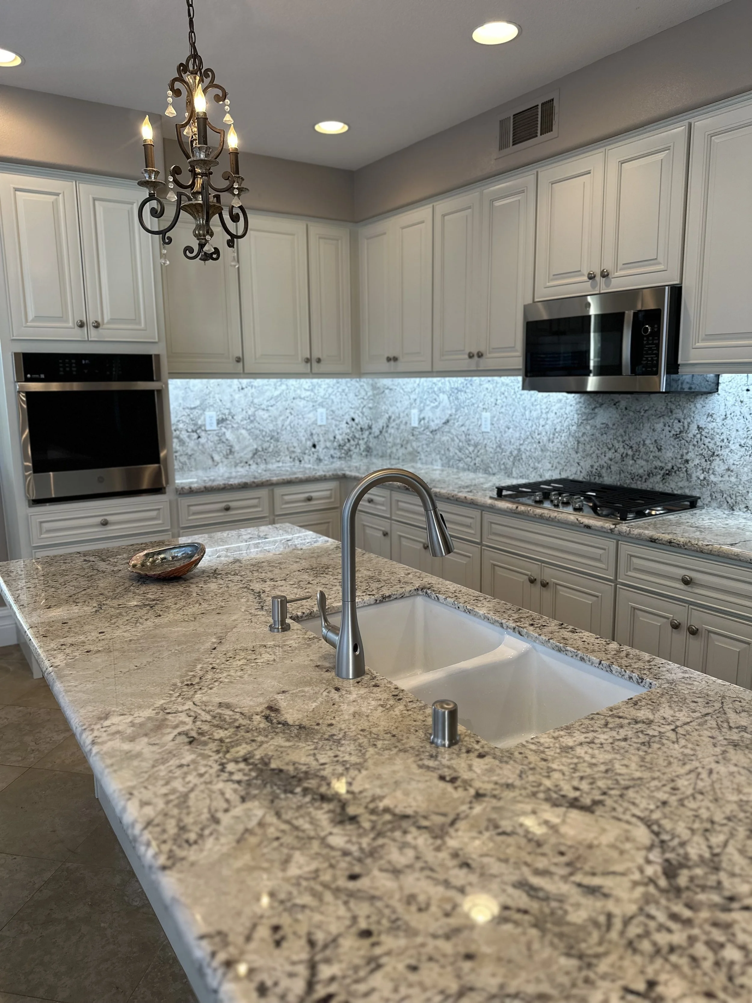 Modern kitchen with granite countertops, white cabinets, stainless steel microwave, oven, and gas stove. There is a double sink with a gooseneck faucet and a decorative chandelier hanging above the island. Multiple recessed ceiling lights are also vi