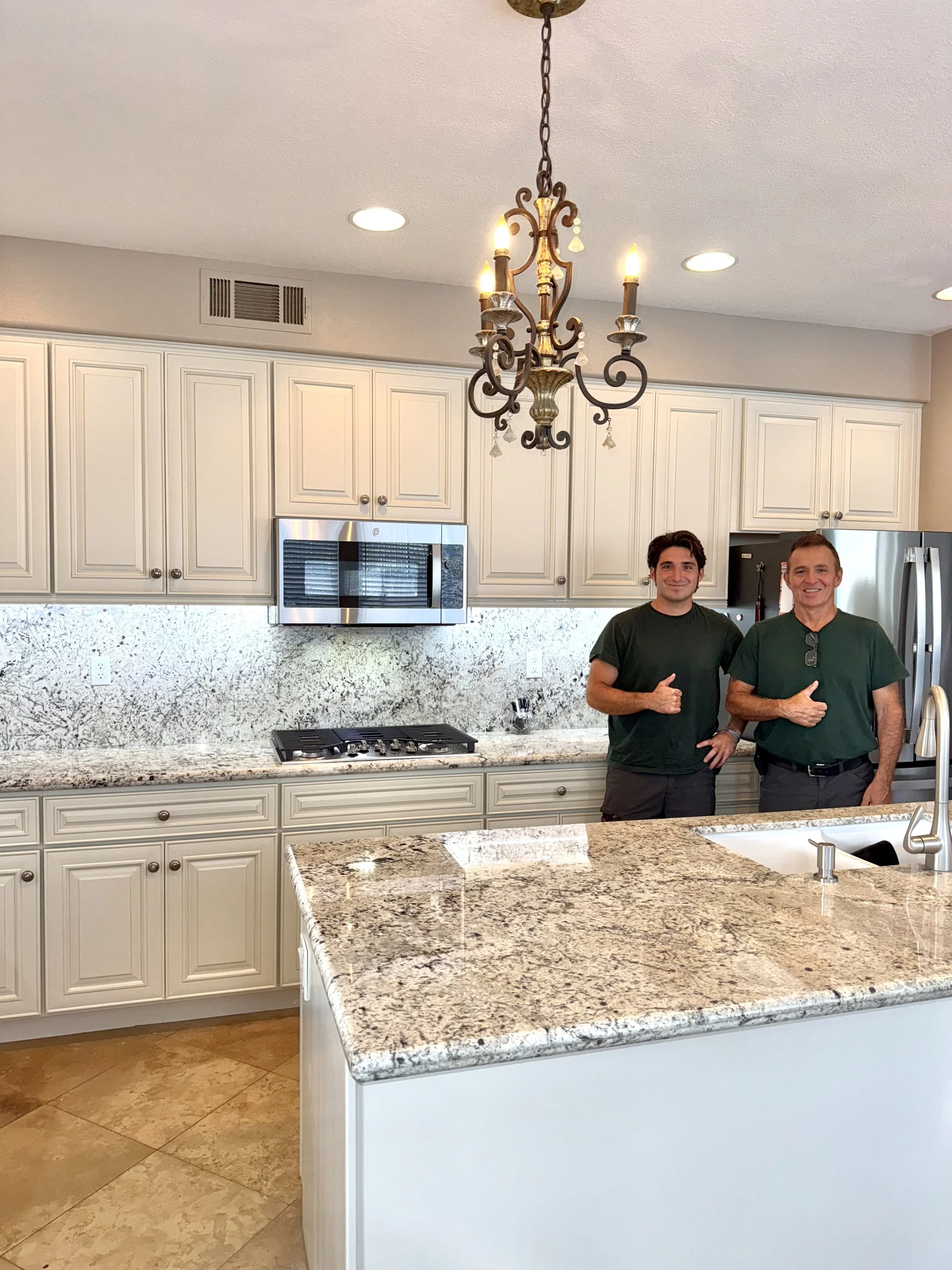 Two men standing in a kitchen, smiling and giving thumbs-up. The kitchen features white cabinet doors, a granite countertop, and a chandelier hanging from the ceiling.