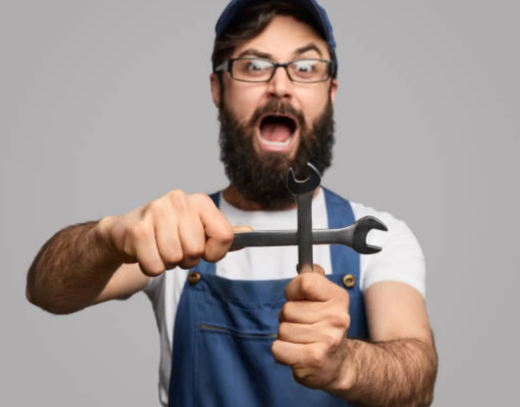 A man with a beard and glasses wearing a blue cap, overalls, and a white t-shirt, holding two wrenches and looking angry.