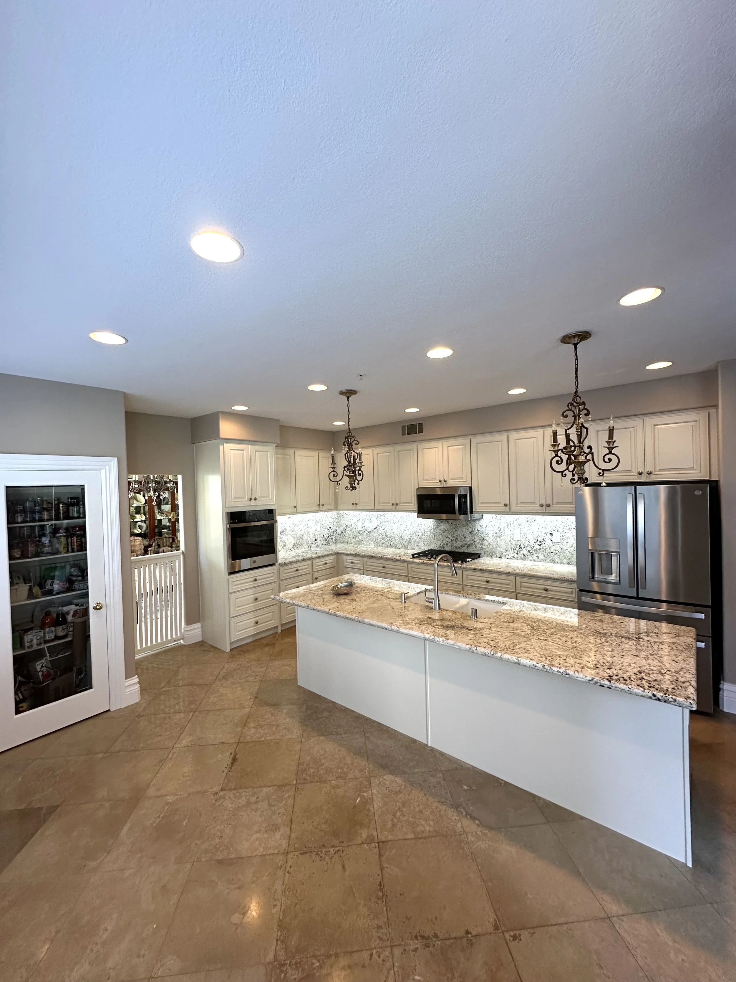 Coffee-colored tiled kitchen floor, white kitchen cabinets, granite countertops, stainless steel refrigerator, built-in oven, microwave, chandelier fixtures, and a kitchen island with a sink.