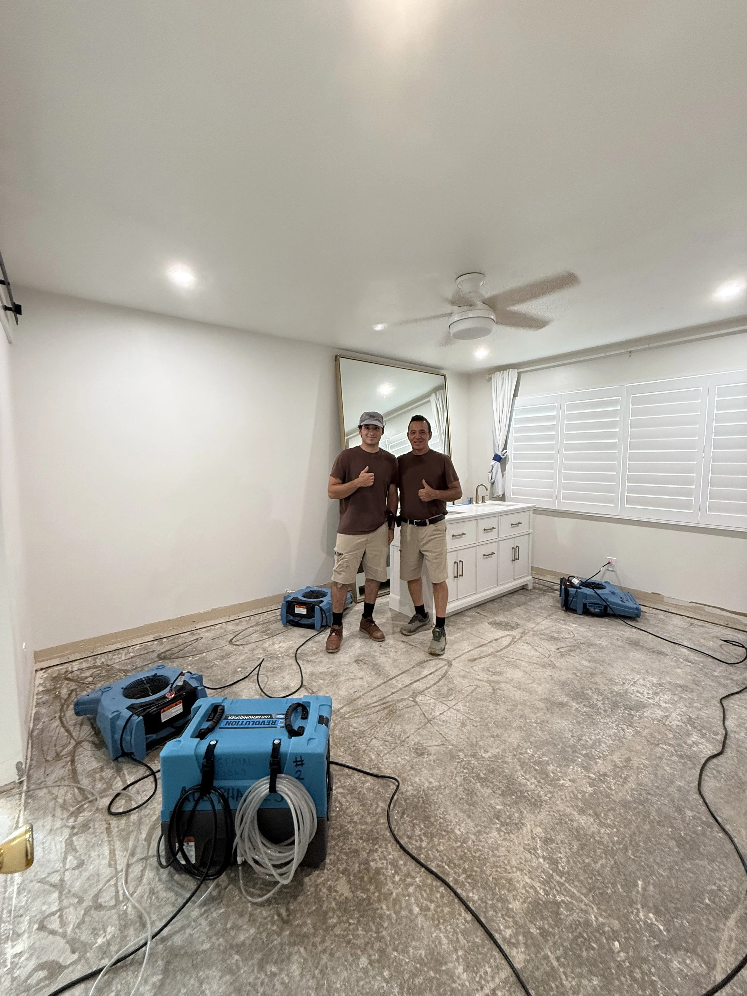 Two men standing in an unfinished room with exposed flooring, surrounded by cooling or dehumidification equipment, giving thumbs up and smiling.