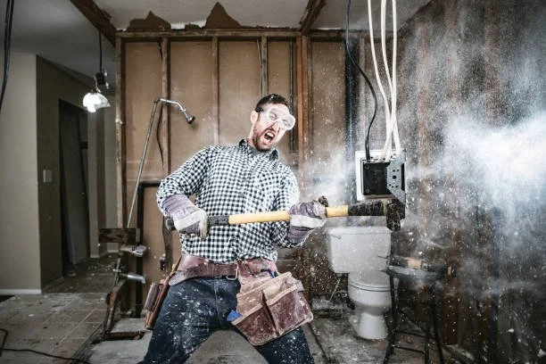 Man in checkered shirt and safety glasses using a sledgehammer on a wall, causing dust and debris, in a renovation or demolition setting.