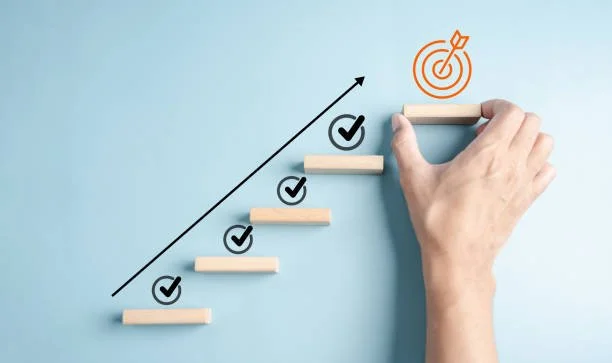 A hand placing a wooden step on a staircase illustration with checkmarks, leading to a target symbol at the top, representing achievement or goal setting.
