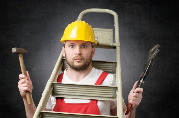 Man wearing a yellow hard hat and red overalls holding a hammer and a wrench, standing behind a ladder.