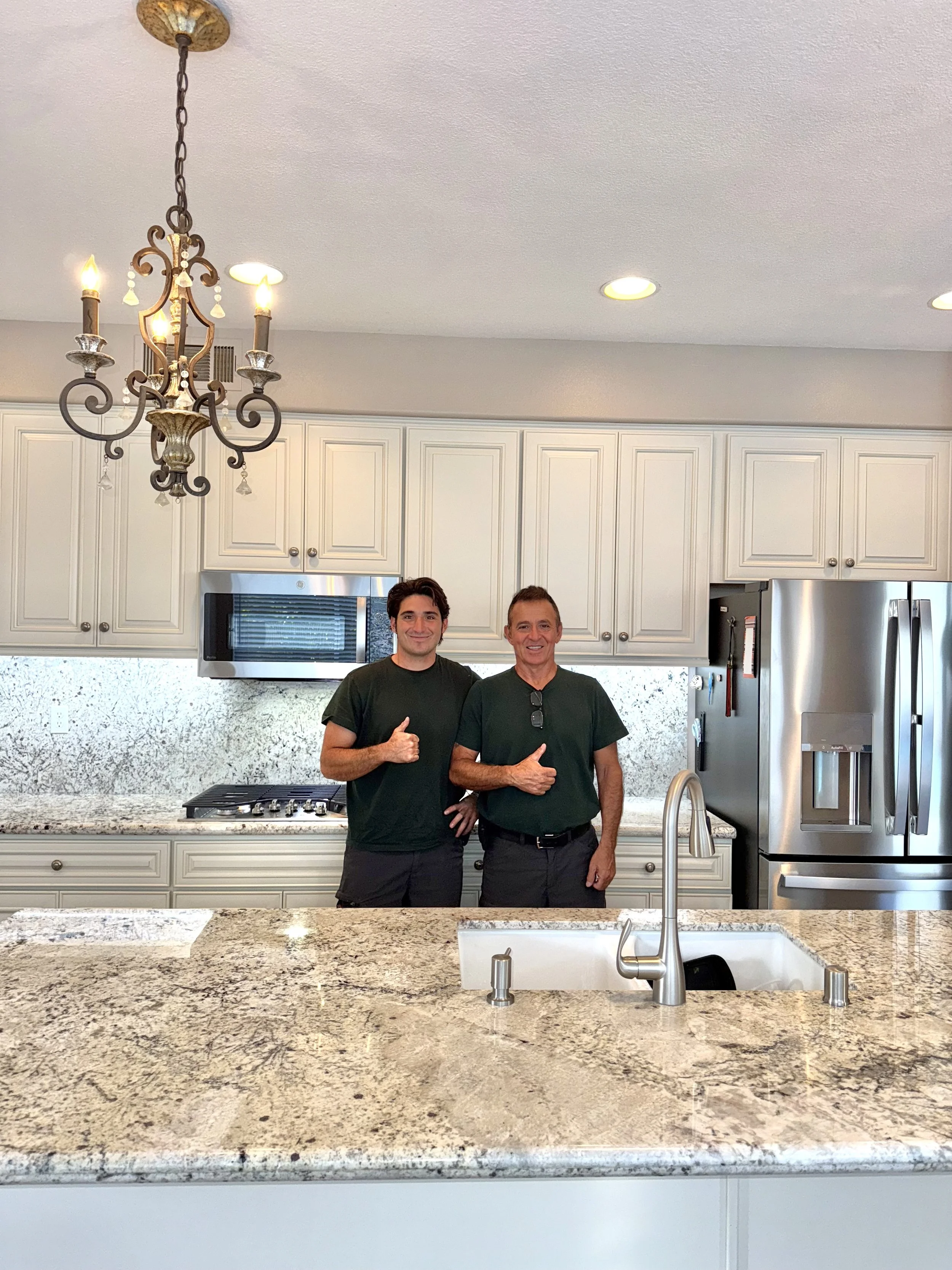 Two men standing in a modern kitchen, giving thumbs-up gestures. The kitchen features white cabinets, a granite counter, a stainless steel refrigerator, stove, and microwave, with a chandelier hanging from the ceiling.