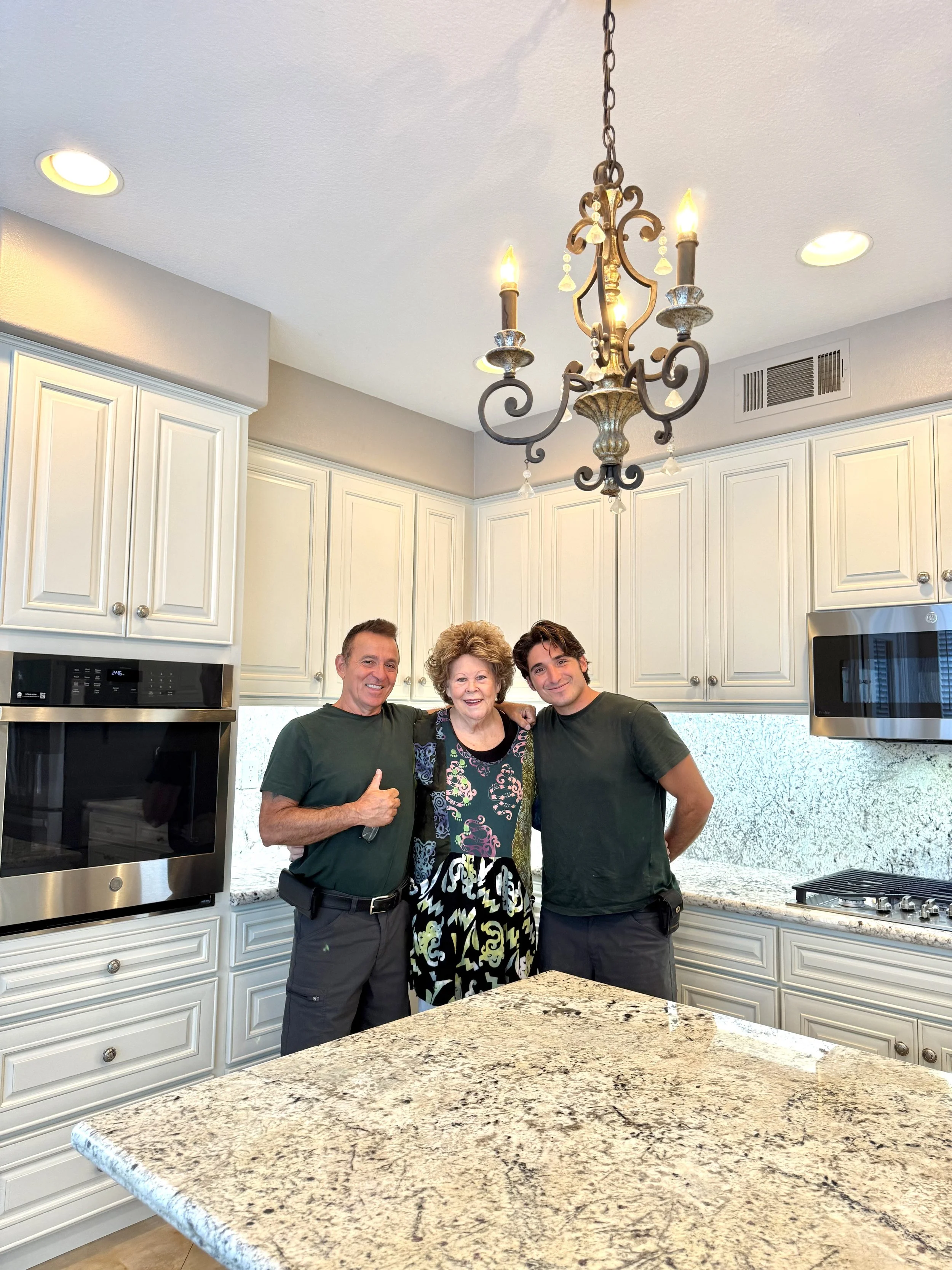 Three people smiling and standing together in a kitchen with white cabinets and granite countertops, with a decorative chandelier hanging from the ceiling.