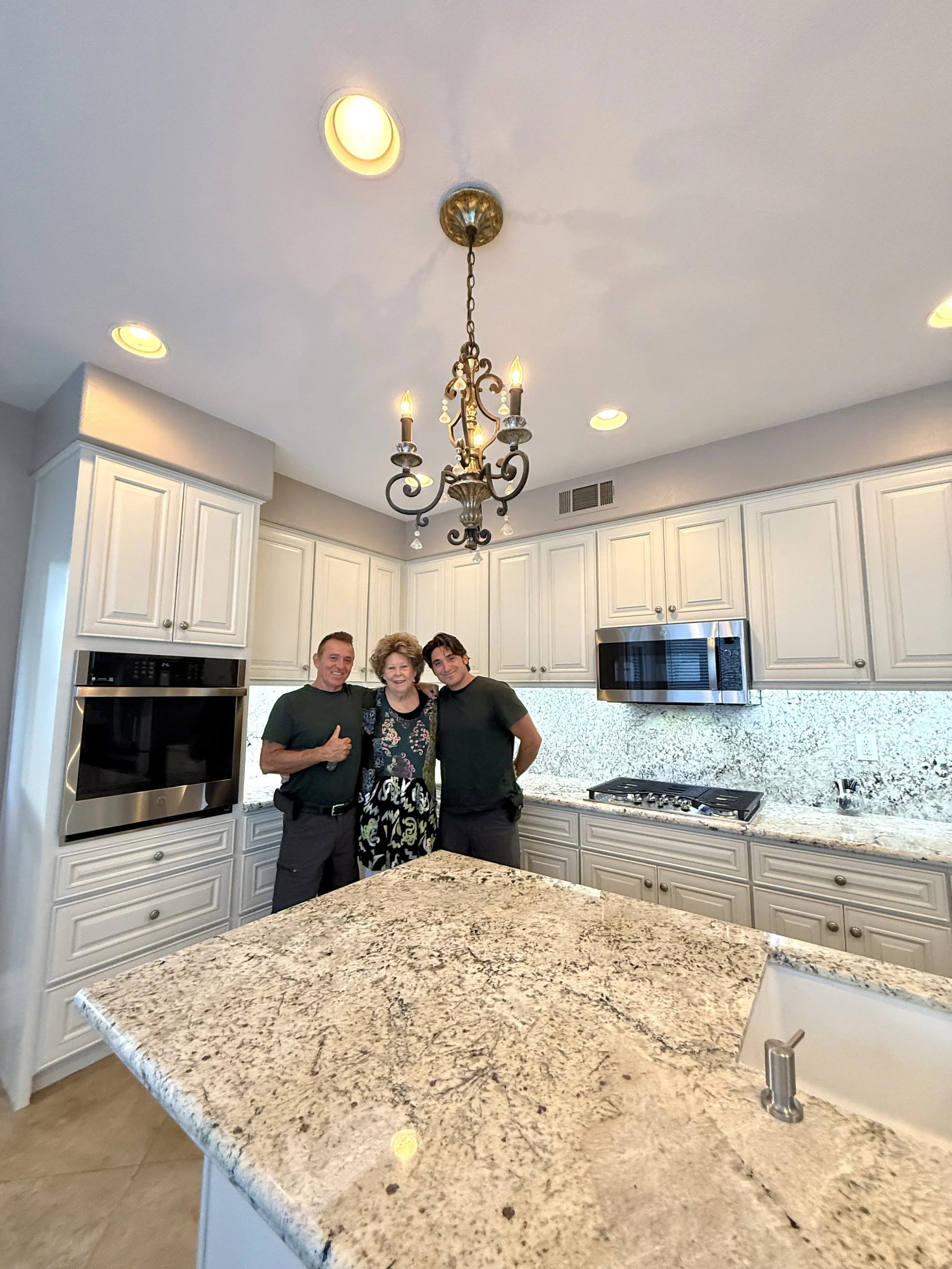 Three people standing together in a kitchen with white cabinets, granite countertops, and a chandelier hanging from the ceiling.
