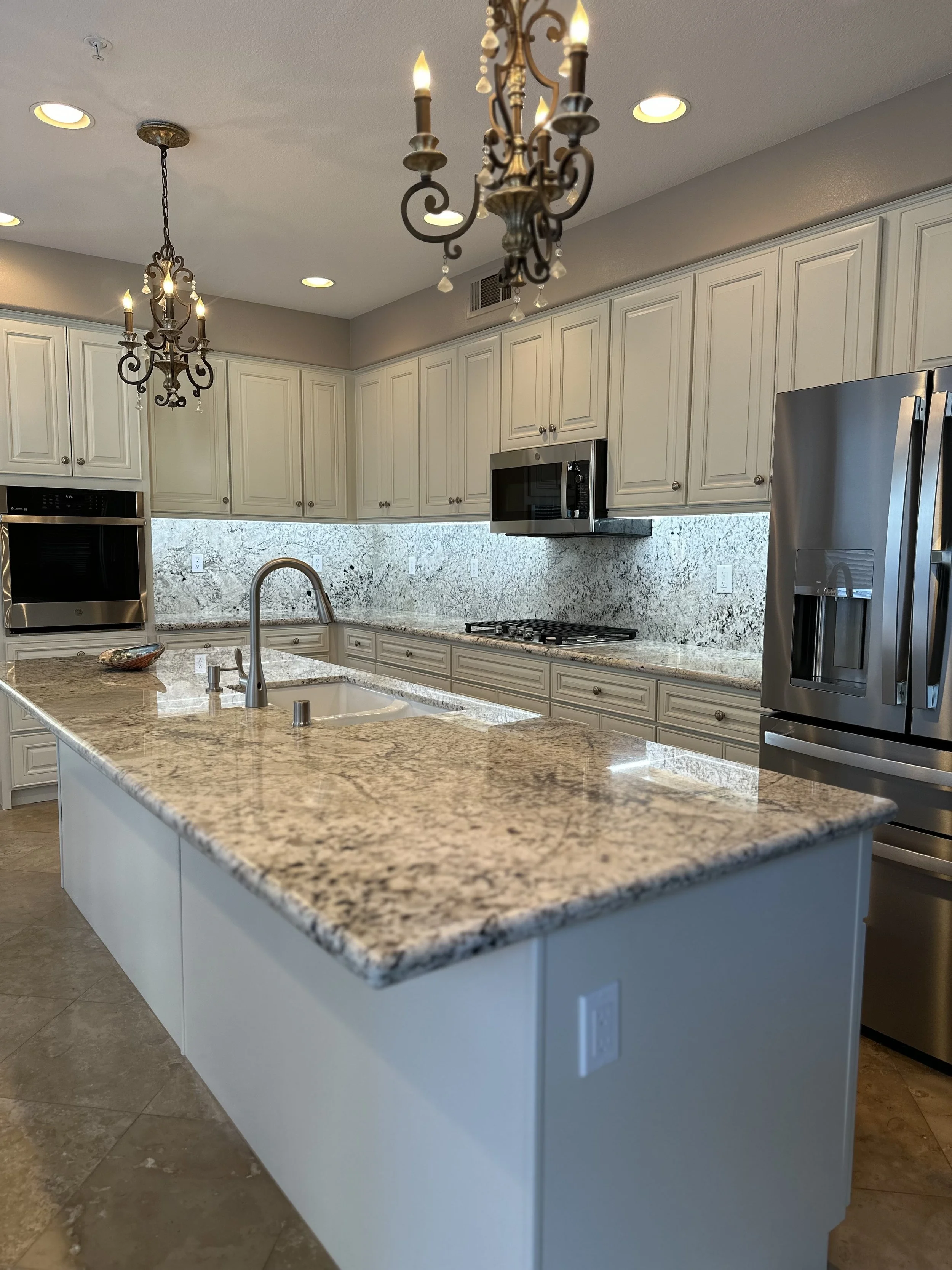 Modern kitchen featuring white cabinets, a granite countertop island with a sink, stainless steel appliances including a refrigerator, oven, microwave, and stove, granite backsplash, and decorative chandeliers hanging from the ceiling.