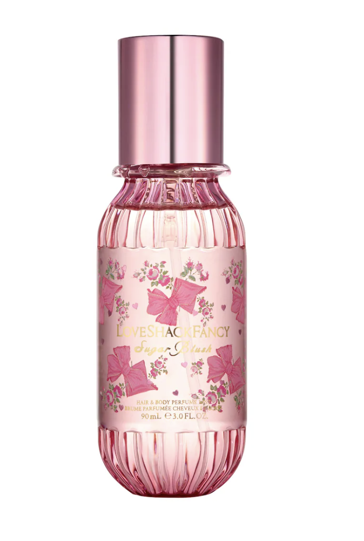 A decorative pink bottle of LoveShackFancy Sugar Blush hair and body perfume mist, featuring a floral rose pattern and a metallic pink cap on a transparent background.