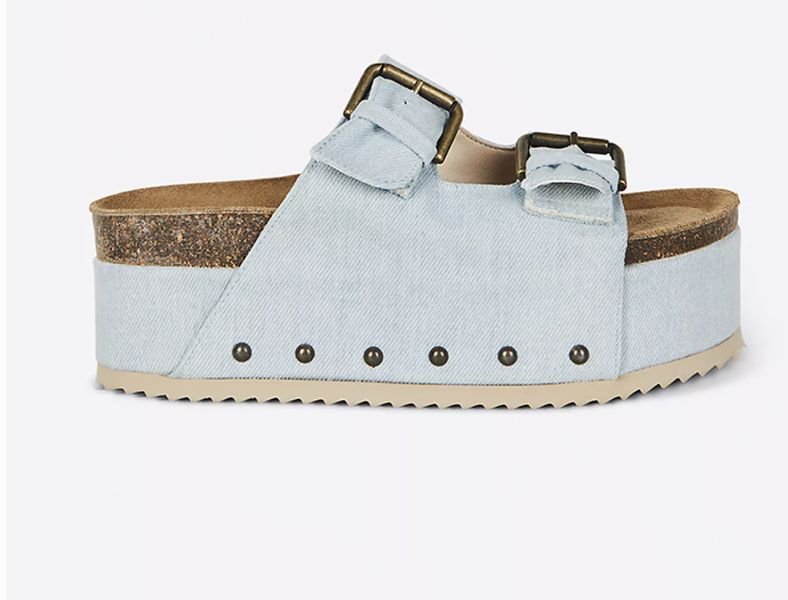 A close-up product shot of a light-wash denim slide clog featuring a thick platform sole, two adjustable straps with antique brass buckles, and small metal stud detailing along the base.