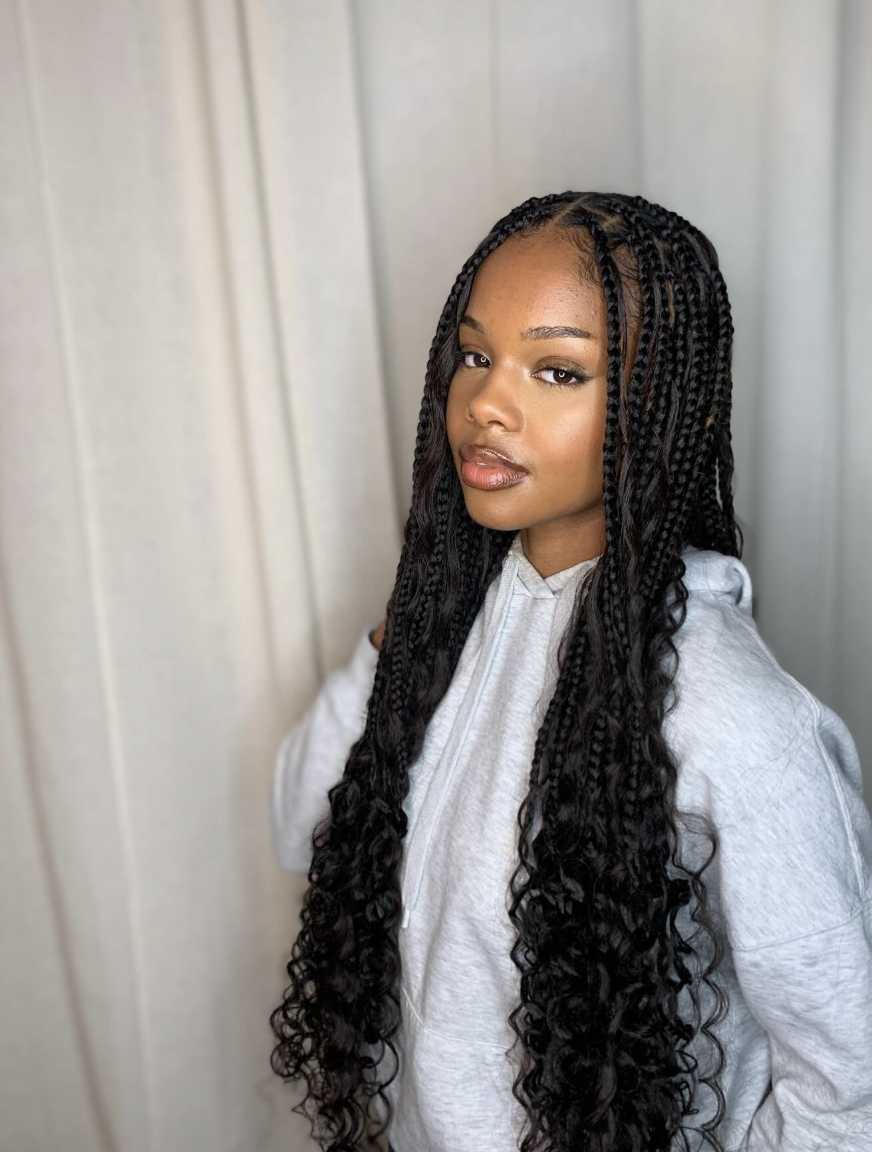 A woman wearing a white hoodie and black jacket, showcasing long goddess braids with curly textured ends that add volume below the shoulders to balance a heart-shaped face.