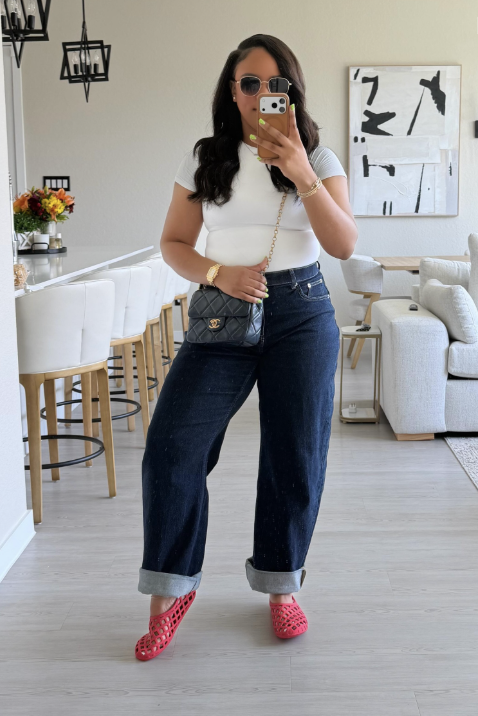 A mirror selfie of a woman wearing high-waisted dark wash straight-leg jeans, a tucked-in fitted white t-shirt, a black quilted crossbody bag, and red jelly flat shoes.