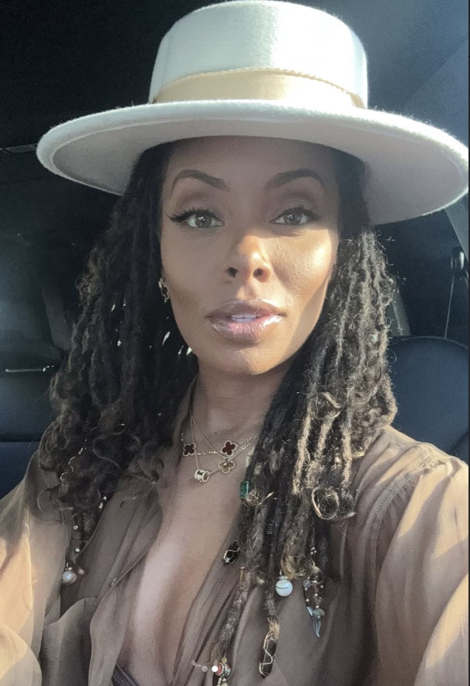 Eva Marcille wearing a wide-brimmed light gray hat and layered gold necklaces, featuring long textured locs with a middle part to show how vertical lines elongate a round face.