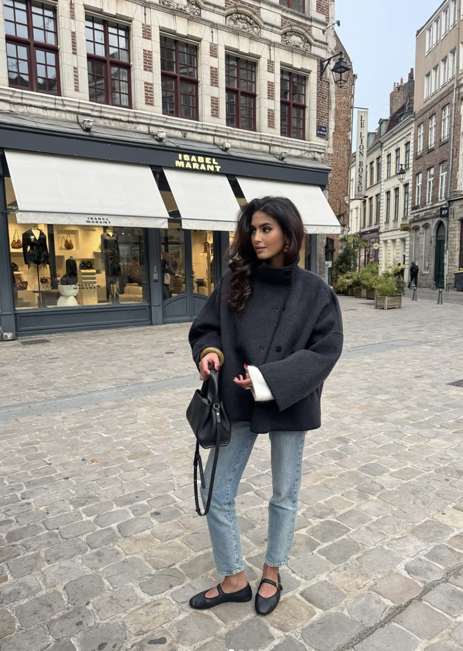 A woman wearing black pointed-toe ballet flats with light wash slim-straight jeans and a dark oversized coat, walking on a cobblestone street.