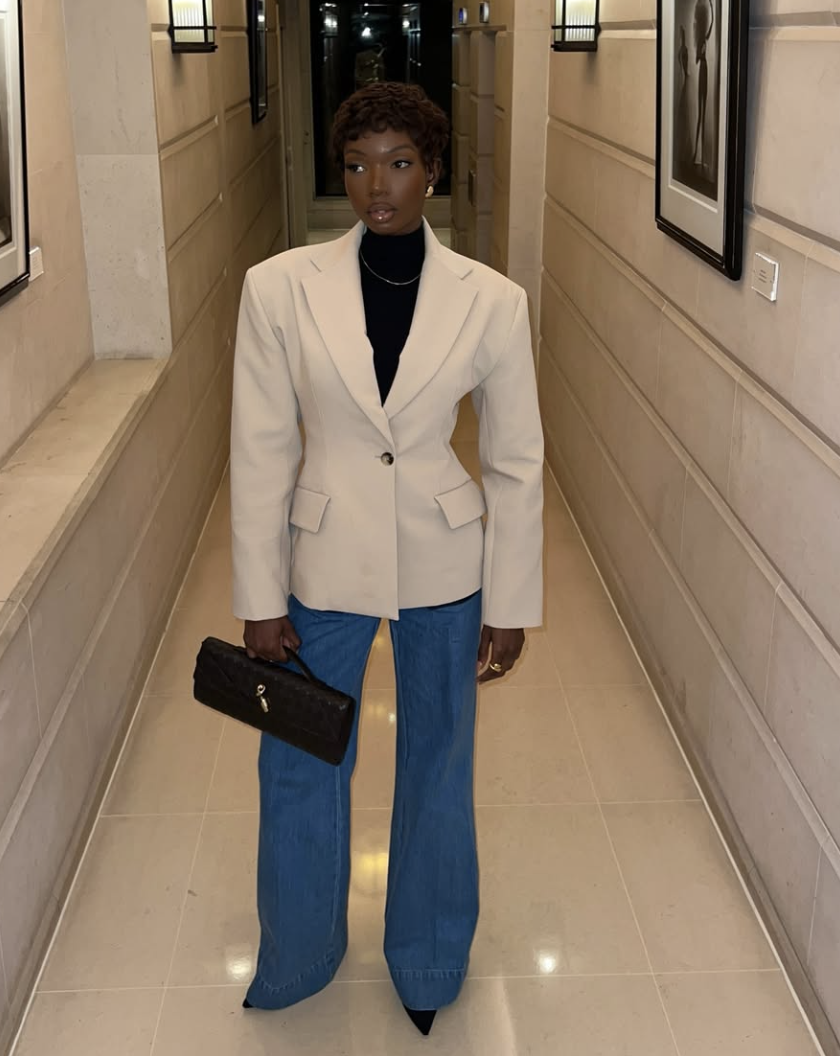 Fashion influencer Sia Luxe wearing a cream nipped-waist hourglass blazer over a black turtleneck and blue bootcut jeans.