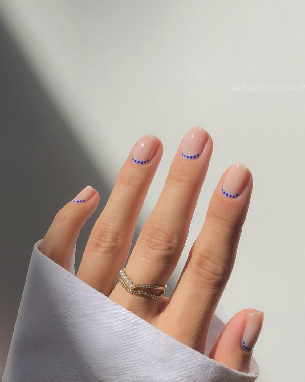 Medium almond-shaped nails with a glossy nude base coat featuring a neat curved line of tiny royal blue dots along the cuticles.