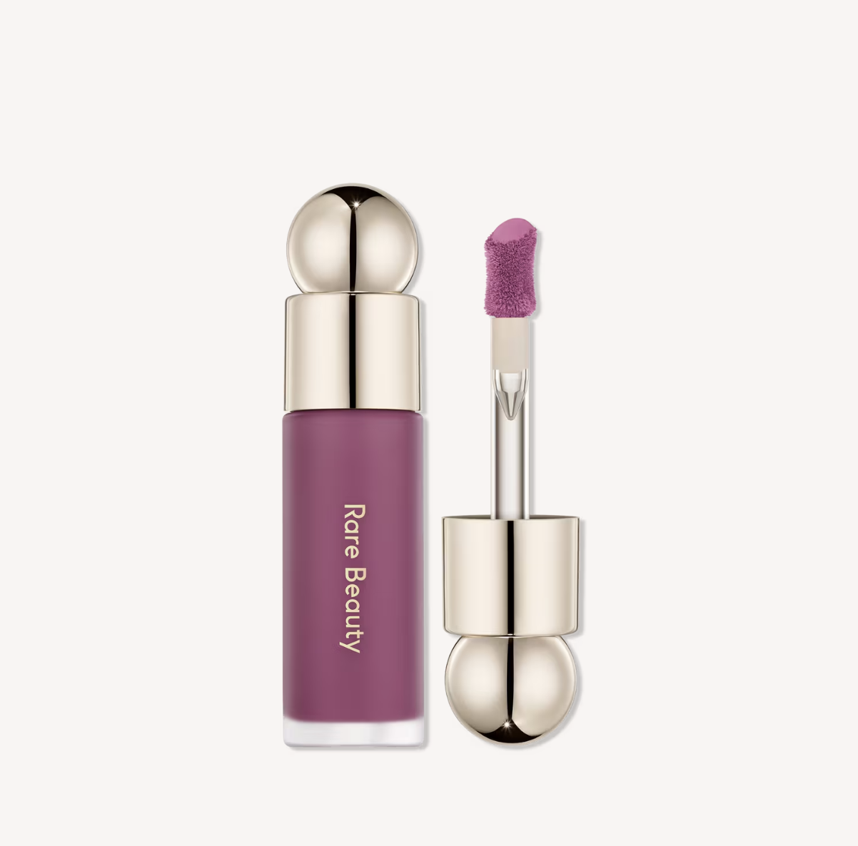 A close-up product shot of the Rare Beauty Soft Pinch Liquid Blush bottle in a vibrant berry-pink shade, featuring its signature gold circular cap and flat-bottomed applicator wand pulled out.