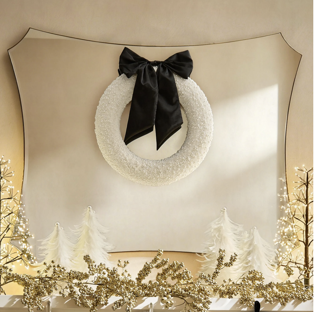 White beaded wreath with a black velvet bow on a scalloped mirror above a mantel with gold berry garland and white feather trees.