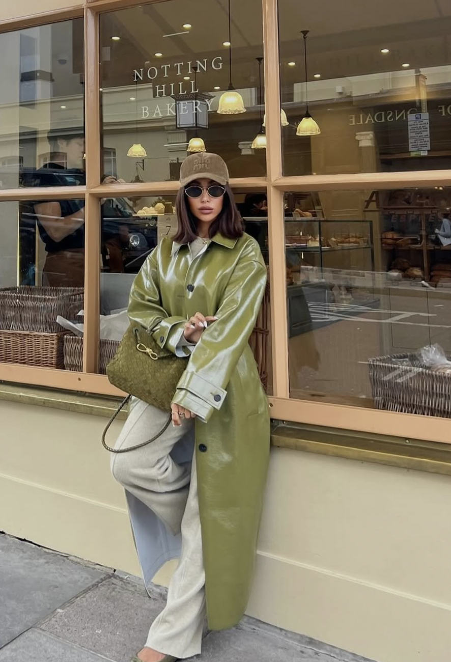 A woman wearing a long, shiny olive green trench coat over grey trousers, styled with a brown baseball cap and a green textured handbag.