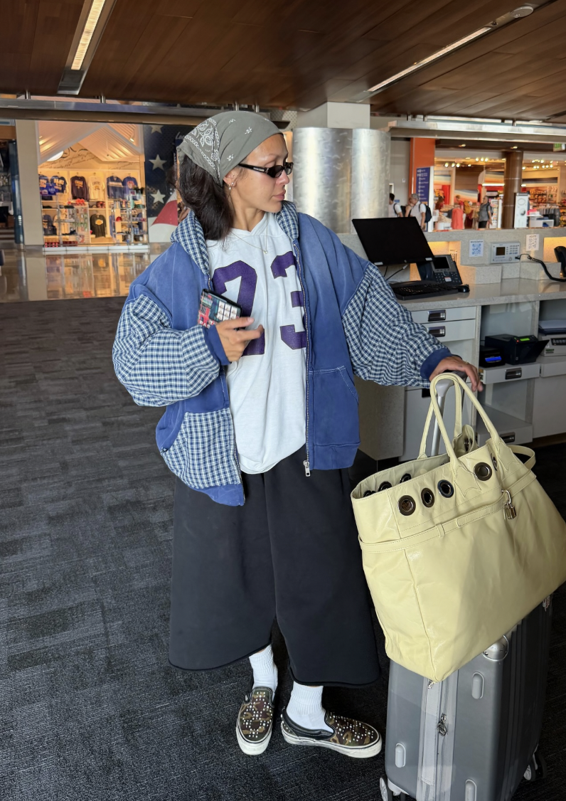 Blue patterned jacket over a numbered jersey with dark knee-length bottoms, covered lower legs, white socks, patterned loafers and a pale-yellow tote.