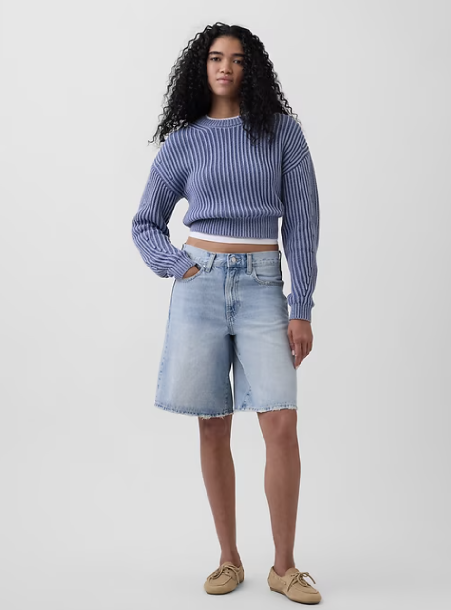 A woman wearing light-wash Gap 11-inch mid-rise longline denim Bermuda shorts with a raw hem, styled with a blue striped sweater and tan boat shoes.