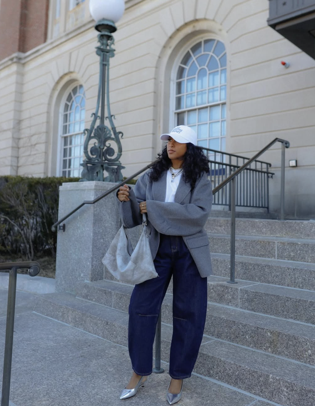 A woman wearing an oversized grey blazer, white t-shirt, dark wash barrel jeans, metallic silver pointed heels, a white baseball cap, and a slouchy grey tote bag.