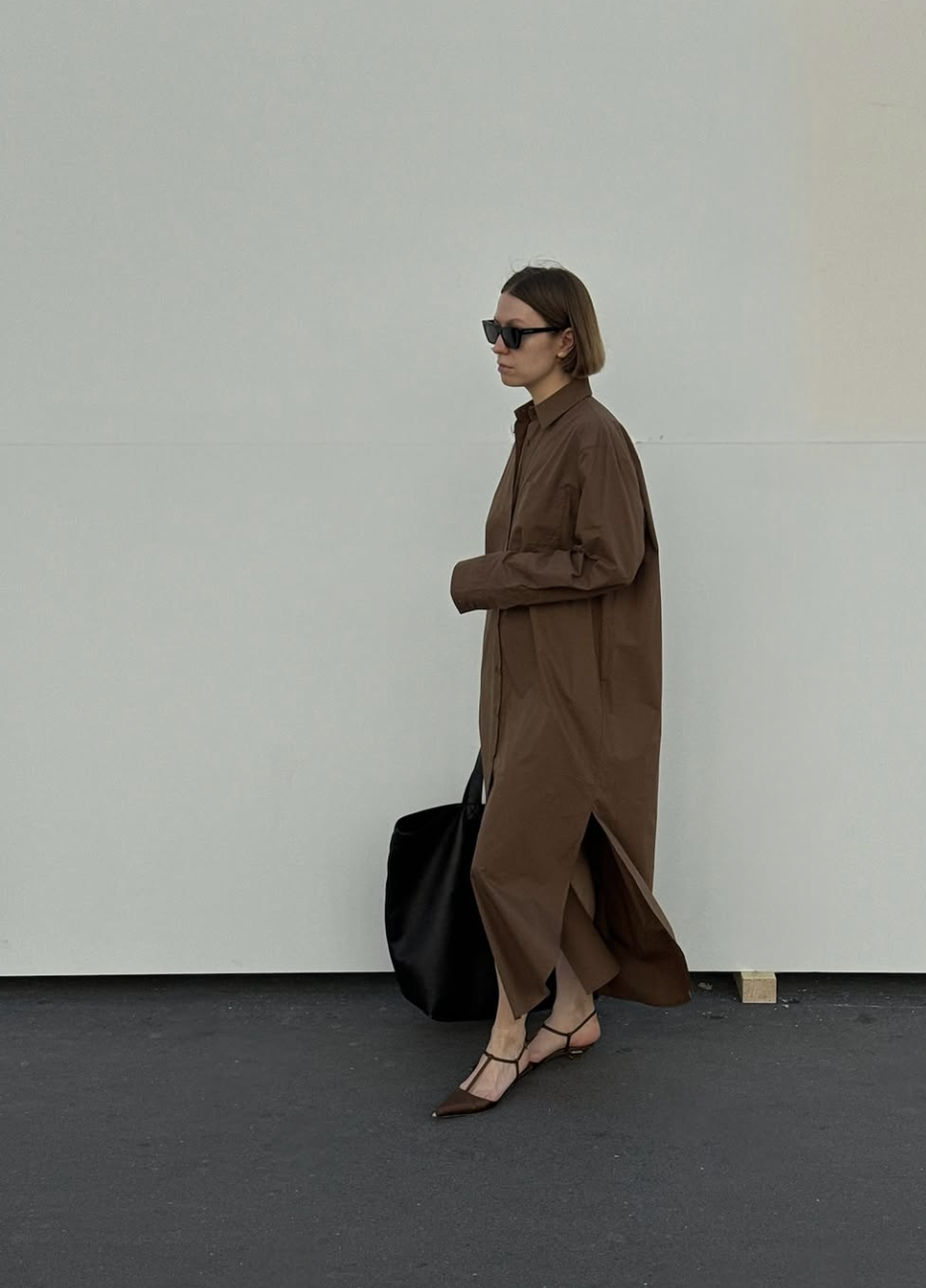 A woman walking in a long chocolate brown oversized shirt dress paired with black pointed-toe T-strap flats and a large black tote bag.  8. Wrap Dress + Slim Ankle Boot