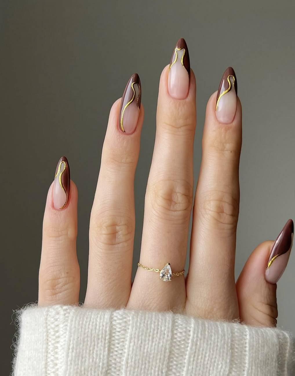 Long almond-shaped gel nails with a latte brown swirl design and 3D metallic gold line art.