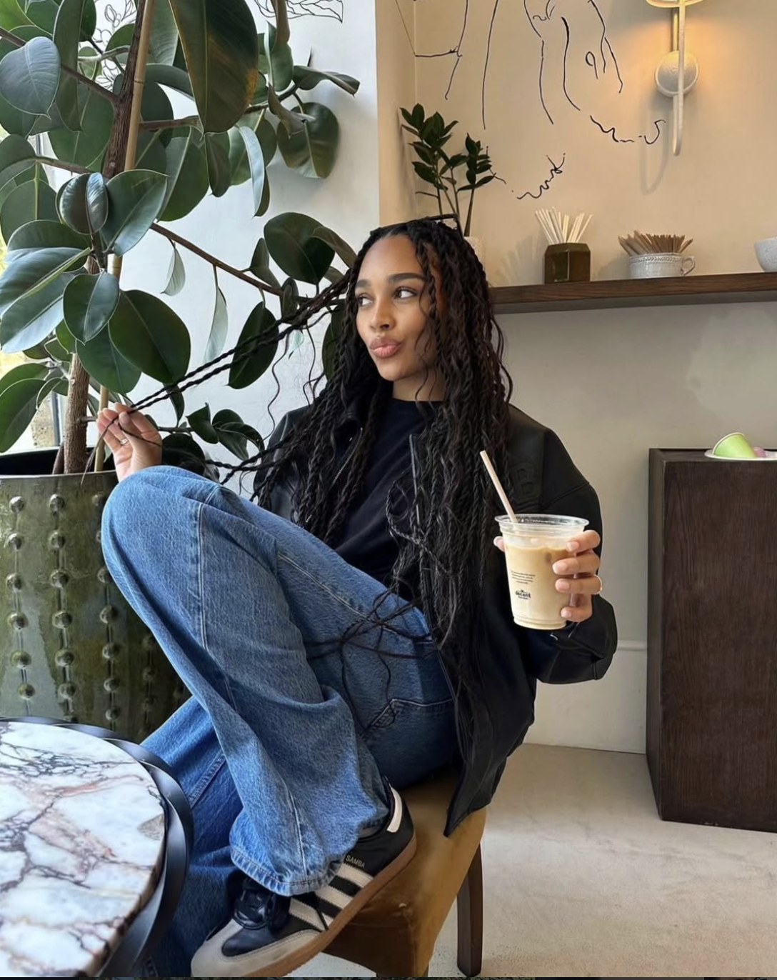 Digital creator Gigi Vassallo sitting at a cafe table wearing a black leather jacket, wide-leg blue jeans, and black Adidas Samba sneakers, holding an iced coffee.