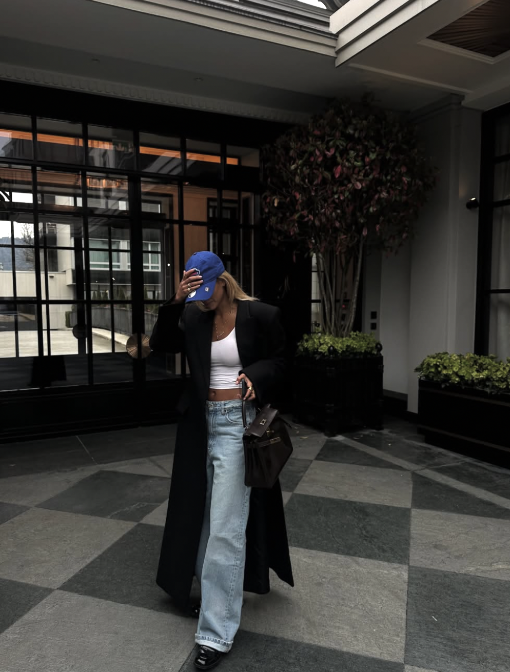 A woman wearing a white cropped tank top, light wash jeans, and a long black coat, accessorized with a blue baseball cap and a brown leather handbag.