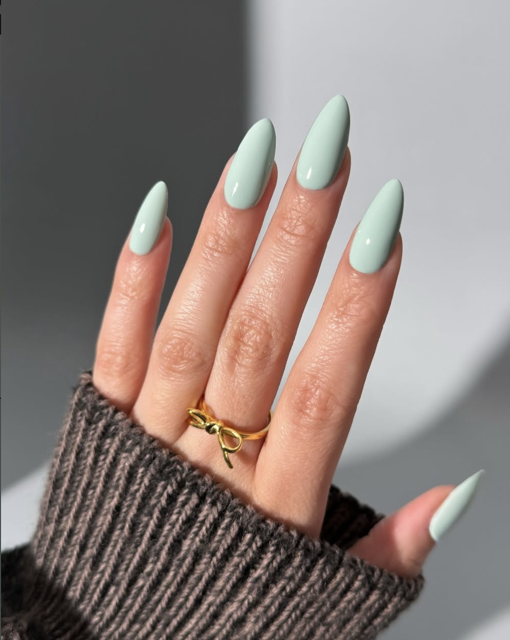 Close-up of long, square-shaped nails with a smooth, opaque mint seafoam green polish, accented by a chunky gold bow ring on the ring finger.