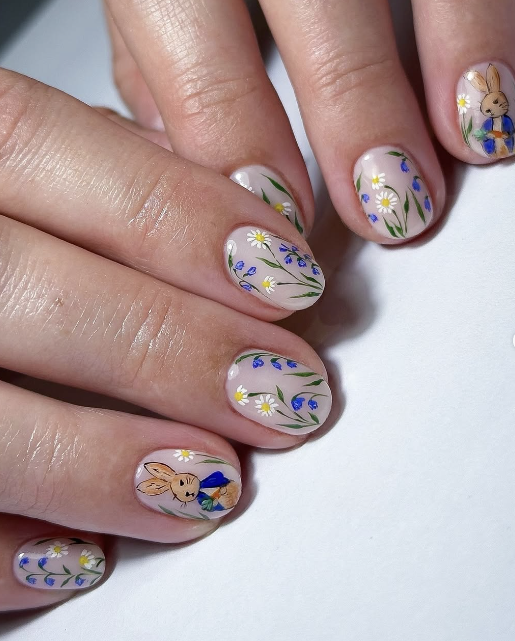 A mismatched spring manicure on short almond nails featuring butter yellow polish, hand-painted carrots, delicate blue and white florals, and a matte speckled "mini egg" texture.