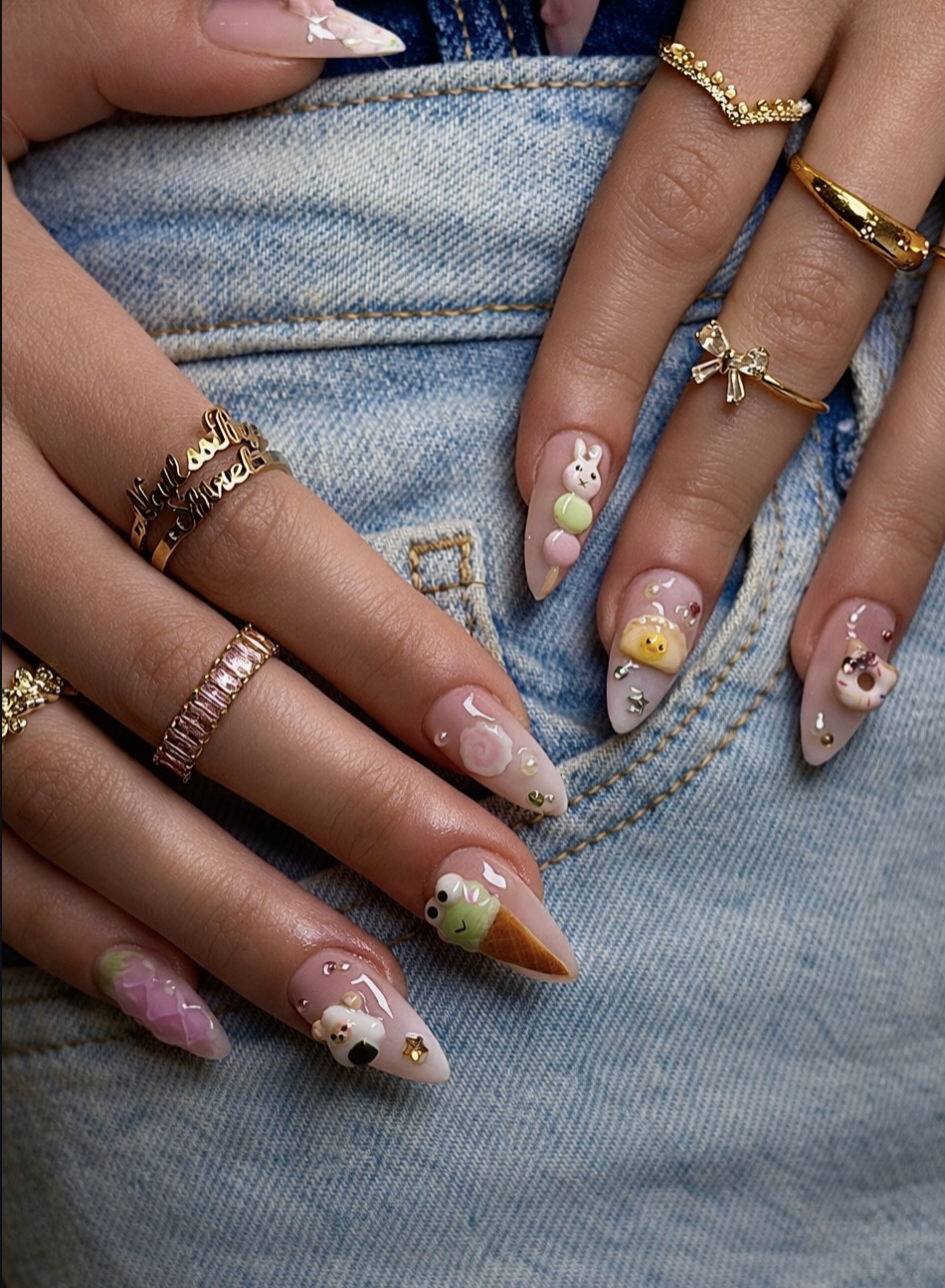 Long almond-shaped nails with a sheer nude base featuring various 3D charms, including a bunny, a frog ice cream cone, a bear onigiri, and colorful pastel candy drops.
