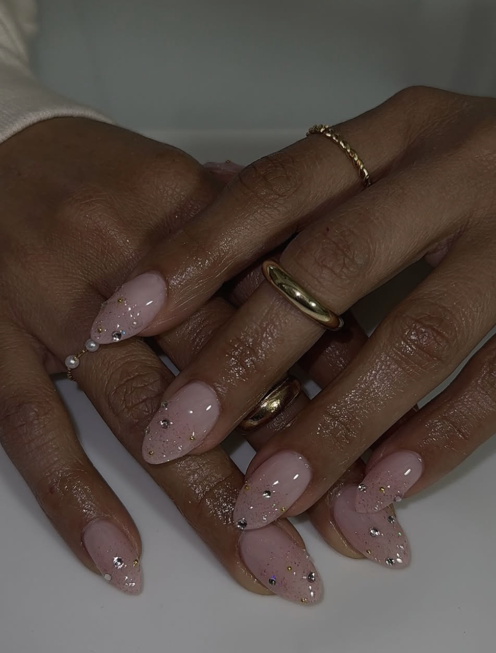 Long almond shaped nails with a sheer pink base, holographic glitter gradient, and small rhinestone accents near the cuticle.