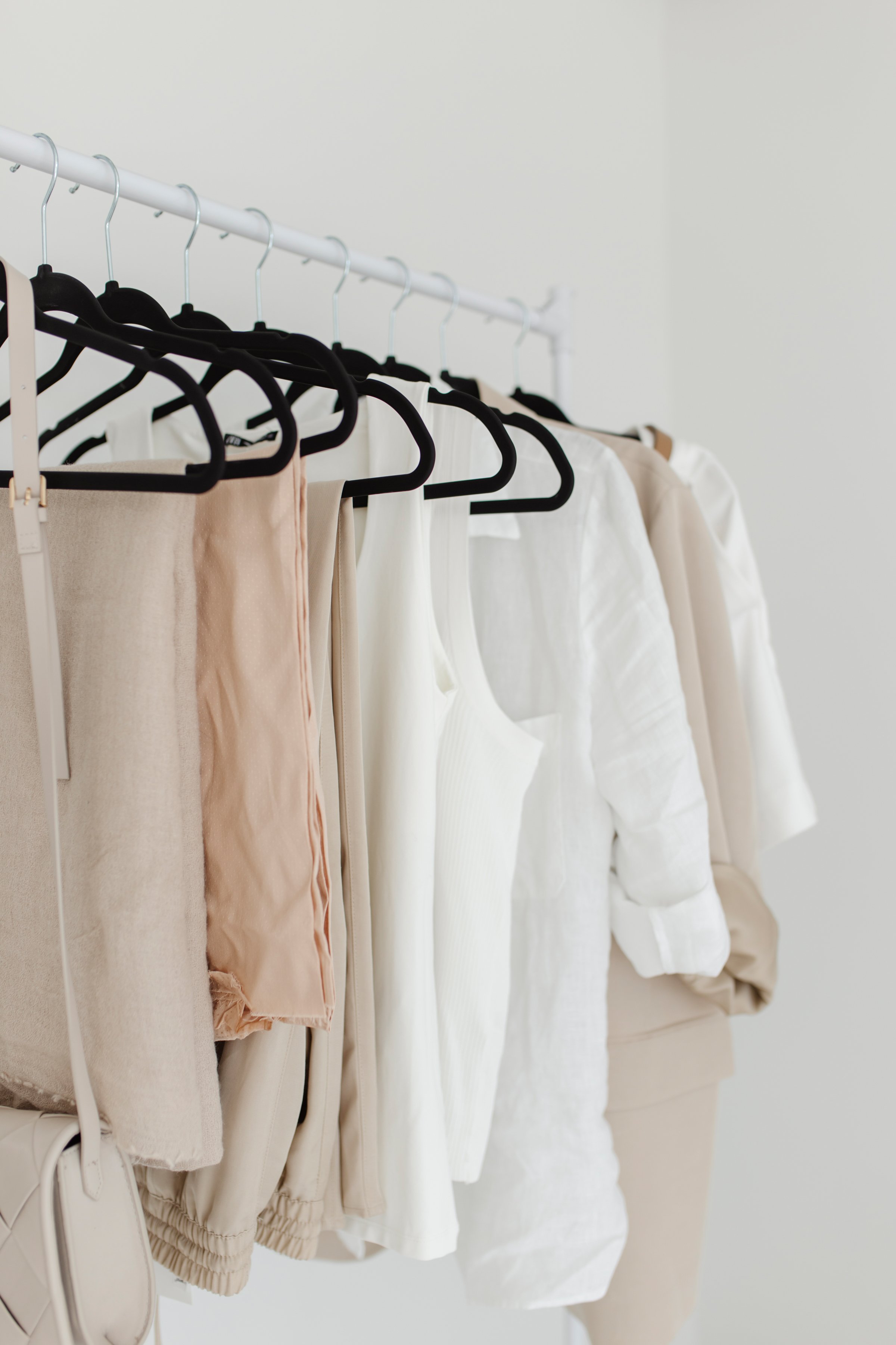 A clothing rack with beige, white, and cream-colored garments on black hangers, including pants, tops, and jackets, against a plain light background.