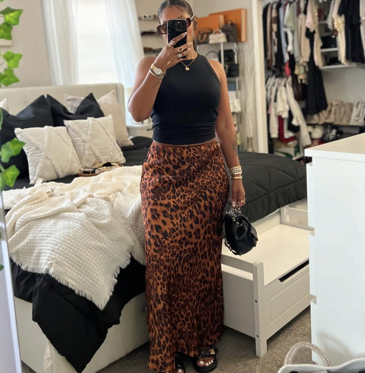 A woman taking a mirror selfie wearing a black sleeveless halter top, a high-waisted leopard print satin maxi skirt, gold bracelets, and holding a small black handbag.
