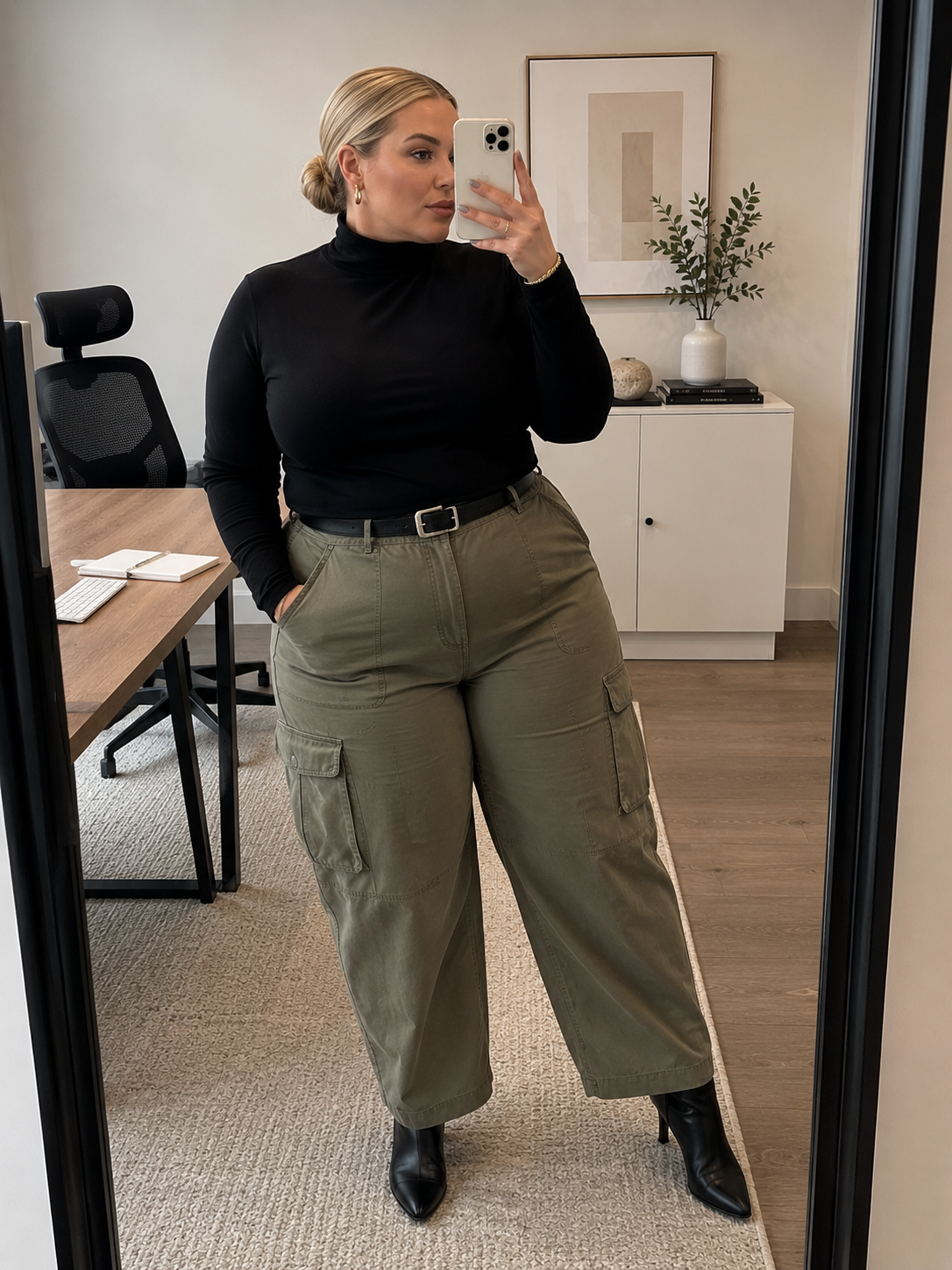 A close-up of a person wearing high-waisted olive green cargo pants with a black leather belt, tucked-in black turtleneck, and black pointed-toe boots.