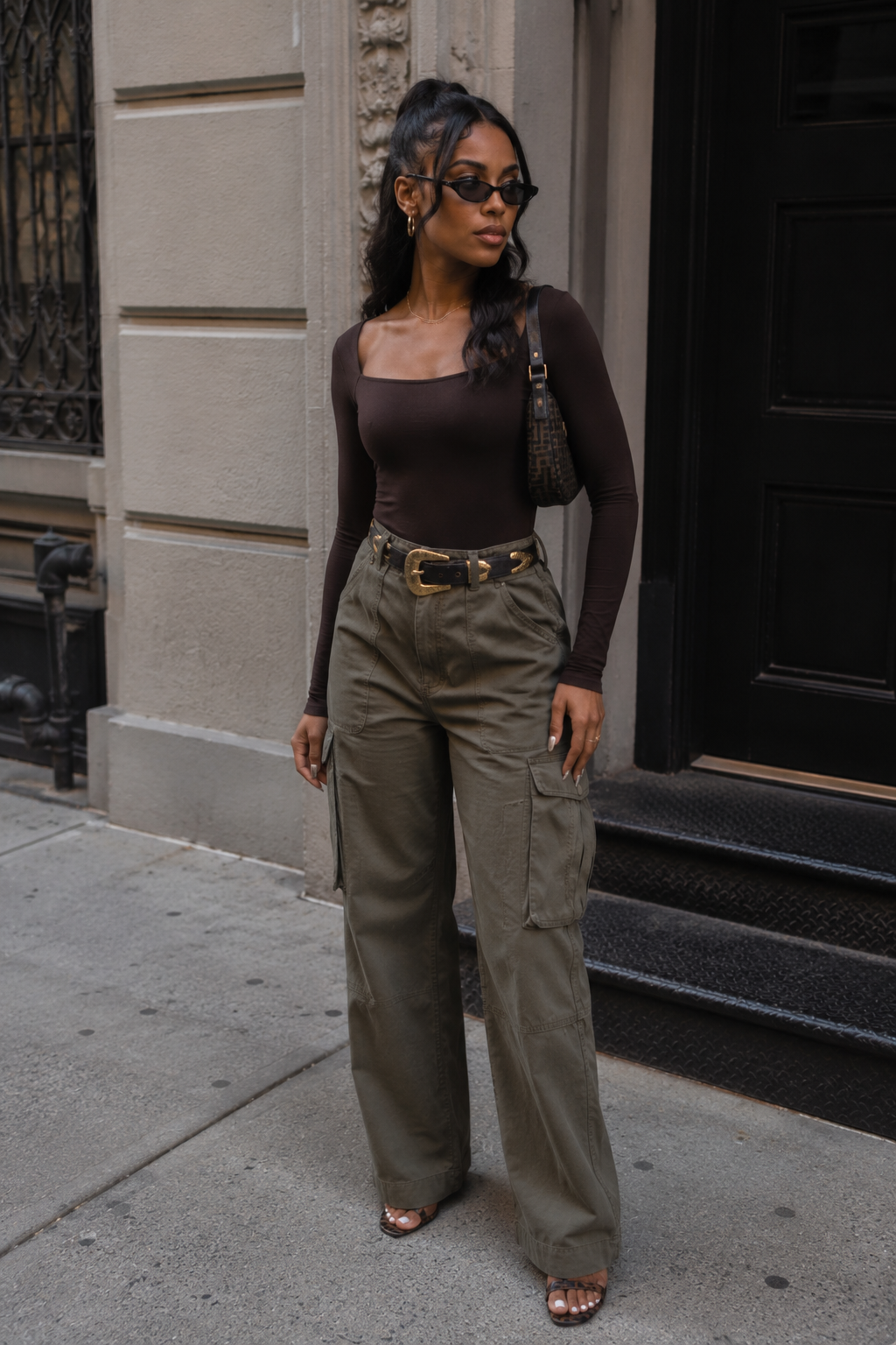 A person wearing a dark brown long-sleeve square-neck bodysuit tucked into olive green wide-leg cargo pants with a gold statement belt and dark sunglasses.