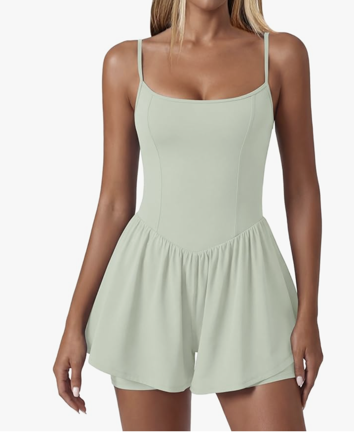 A woman wearing a casual light sage green romper featuring a scoop neckline, thin spaghetti straps, a defined smocked waistline, and relaxed, wide-leg shorts.